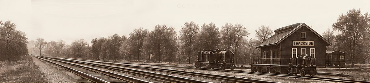 Sepia-toned vintage photograph of a small train station building beside multiple railroad tracks, with trees in the background and a train visible on the tracks.